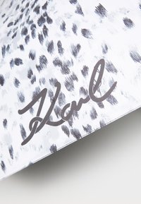 White fabric with black abstract spots features a prominent black handwritten signature "Karl." The texture appears smooth and soft.