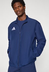 Μπλε ναυτικό μπουφάν με φερμουάρ, κατασκευασμένο από λείο ύφασμα, με γιακά, πλαϊνές τσέπες και λευκό λογότυπο στο αριστερό στήθος.