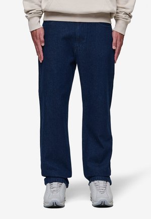 Parte inferior del cuerpo de una persona que lleva vaqueros holgados azul oscuro, sudadera beige y zapatillas blancas, de pie con las manos a los lados.