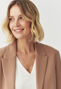 Capelli biondi e ondulati che incorniciano il viso, indossando un blazer beige sopra una maglietta bianca, con una collana a pendente circolare dorata come accessorio principale.