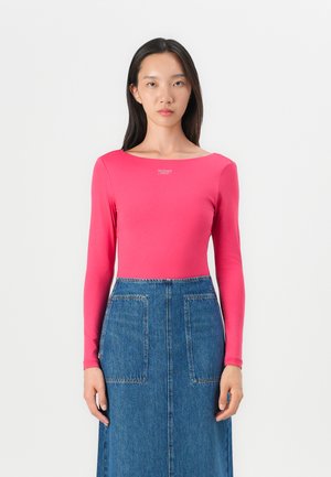 Fitted long-sleeve top in bright pink fabric, scoop neckline, featuring a silver logo accent. Paired with a high-waisted denim skirt.