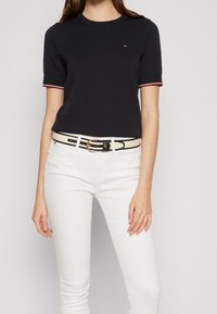 Camiseta de manga corta azul marino con detalles rojos y blancos, combinada con unos jeans blancos ajustados y un cinturón negro con una hebilla dorada.