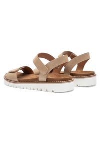 Pár béžových semišových sandál s nastaviteľnými remienkami a bielymi rebrovanými gumovými podrážkami, zobrazený zo zadného trojštvrťového uhla.