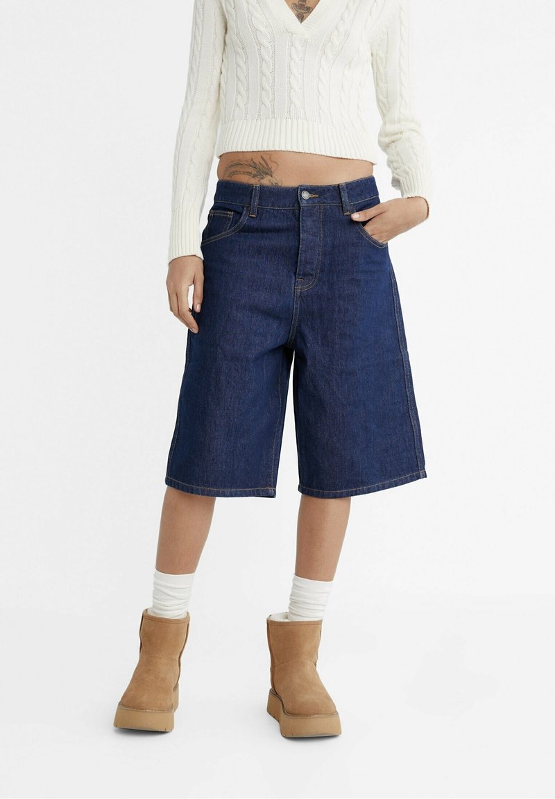 Personne portant un short bermuda en denim bleu foncé taille haute, un pull court à torsades blanc, des chaussettes blanches et des bottines marron clair, main dans la poche.