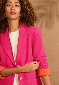 Fuchsiový blazer s upraveným střihem, který má oranžové manžety, dva zlaté knoflíky a texturovanou tkaninu. Model má pod ním krémový top.