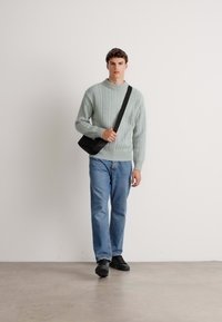 Man walking wearing a light gray ribbed sweater, blue jeans, black sneakers, and a black crossbody bag against a plain white background.