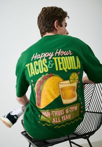 Homme assis sur une chaise en fil métallique noire, portant une chemise verte avec une grande impression au dos représentant un taco, un verre de tequila avec citron vert, et le texte « Happy Hour Tacos & Tequila Lun-Jeu 18h-19h. »