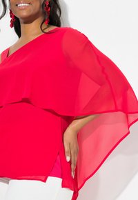 Top rosso trasparente con design a spalla unica, sovrapposto a una base rossa solida, abbinato a pantaloni bianchi. Tessuto liscio, leggero.