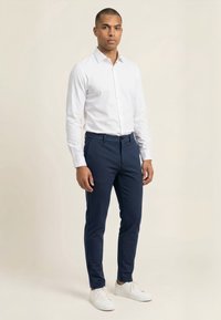 Mand stående iført en hvid skjorte med lange ærmer, navyblå slim-fit bukser og hvide sneakers på en beige baggrund.