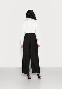 Bílá košile dlouhého rukávu a černé kalhoty s vysokým pasem a širokými nohavicemi, nošeny s černými lodičkami, viděné zepředu. Jednoduché textury, minimalistický design.