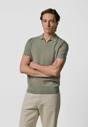 Kortærmet polo i afdæmpet grøn, lavet af strikstof. Har en v-udskæring og ribkant nederst. Bæres med lyse beige bukser.