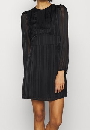 Femme portant une robe mini noire transparente à manches longues avec des rayures verticales et un col montant, debout devant un fond uni.