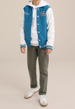 Chaqueta varsity azul y blanca con el número 86 bordado, combinada con una sudadera blanca, pantalones verde oliva y zapatillas beige y blancas.