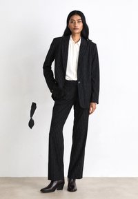 Costum negru cu dungi fine, cu sacou ajustat și pantaloni drepti, purtat cu o cămașă albă. Textură netedă și design structurat. Botine maro până la gleznă.