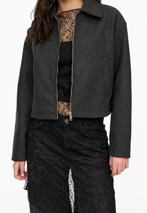 Frau trägt eine dunkelgraue Jacke mit Reißverschluss über einem schwarzen Spitzentop und einem strukturierten schwarzen Spitzenrock mit vorderem Schlitz.