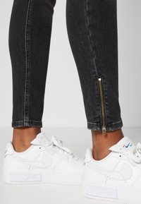 Jeans negros ajustados con detalles de cremallera en el tobillo, combinados con zapatillas bajas blancas que tienen una suela texturizada y un logo multicolor.