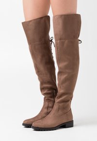 Brązowe zamszowe wysokie botki do uda z okrągłym noskiem, niskim obcasem na słupku, zapięciem na zamek błyskawiczny oraz koronkowym wykończeniem z tyłu. Teksturowana powierzchnia.