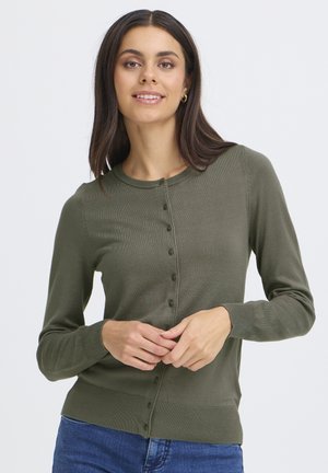 Mladá žena v olivovo zelenej zapínanej vestke a modrých džínsoch, stojaca a usmievajúca sa na jednoduchom bielom pozadí.