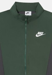 Nike zipzáras dzseki sötétzöld anyagból, magas gallérral, fehér logóval és kontrasztos fekete oldalsó panelekbő. Simaság, elegáns dizájn.