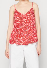 Top sin mangas rojo con lunares blancos, escote en pico, cierre de botones en la parte delantera y dobladillo peplum. Tela ligera con una textura suave.