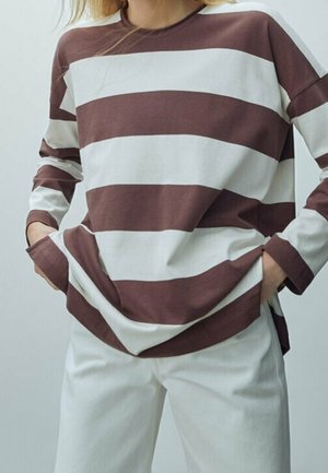 Person wearing long-sleeve brown and white horizontal striped shirt with hands in pockets, paired with white pants against plain background.