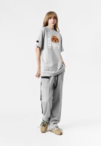 Camiseta gris de gran tamaño con gráfico de un oso y texto "Black", combinada con pantalones de chándal grises con bolsillos laterales y acentos de stripp negra.