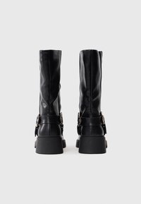 Par de botas de cuero negras a media pantorrilla con tacones gruesos, correas decorativas y hebillas metálicas, mostradas desde atrás sobre un fondo blanco.