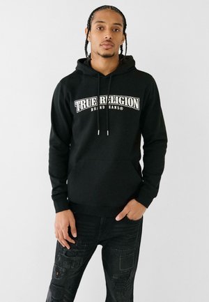 Felpa nera con cappuccio e testo stampato in bianco "TRUE RELIGION" e "BRAND BANS." Abbinata a jeans neri strappati con dettagli a toppa.