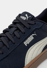 Marinblå mockaskor med platta snören, vit läderrand och brun gummisula. Har Puma-logotypen på tungan och sidan.