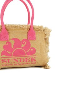 Sac en toile de jute beige avec des poignées roses et un logo en vagues roses. Présente des franges le long du bord supérieur et le texte de la marque en caractères gras.