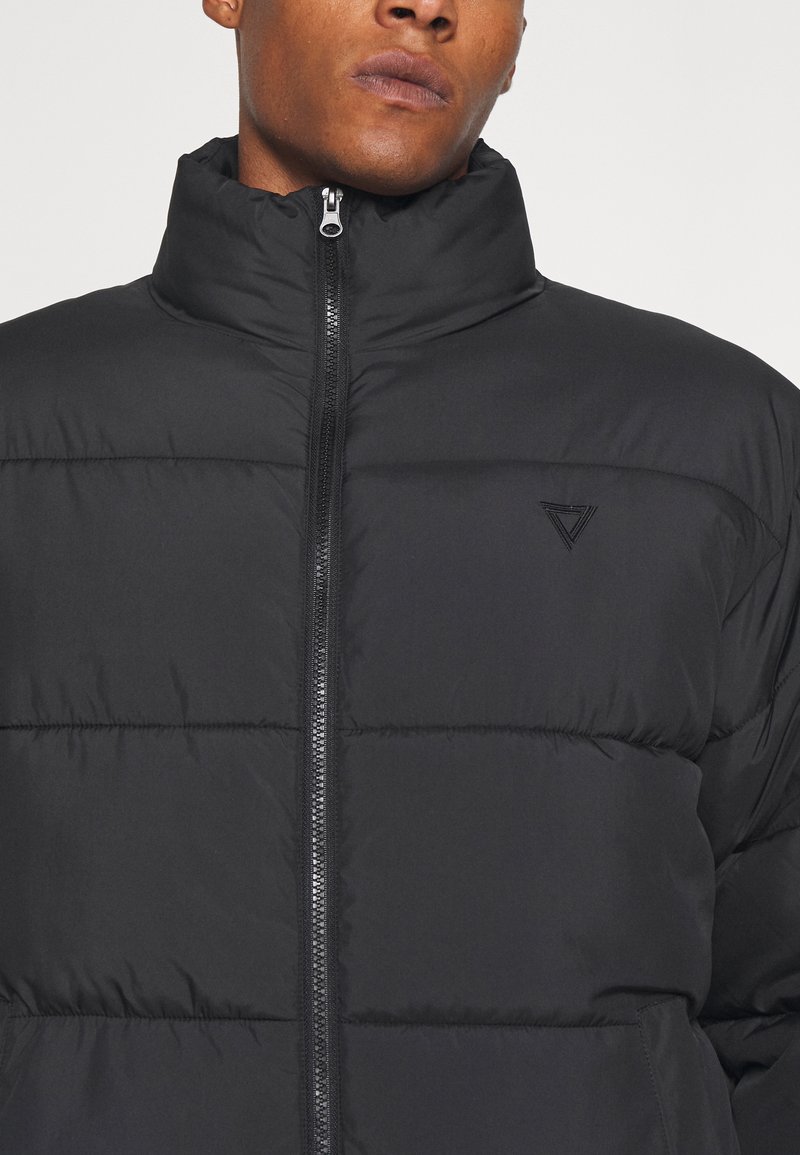 Schwarze Pufferjacke mit hohem Kragen, mit horizontalen Nähten und einem Reißverschluss auf der Vorderseite. Subtales dreieckiges Logo auf der linken Brust.