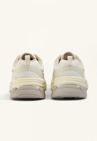 Beige und weiße Sportschuhe mit einem Mesh-Obermaterial, runder Zehenpartie, gepolstertem Kragen und dicker, strukturierter Sohle. Die Rückansicht zeigt das Detail der Zugschlaufe.