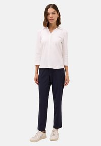 Polo a maniche lunghe bianco con colletto e patta con tre bottoni, abbinato a pantaloni slim-fit blu navy e sneakers beige.