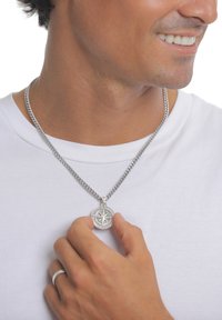 Silberne Halskette mit rundem Kompassanhänger, der feine Details und eine polierte Oberfläche aufweist, getragen über einem weißen T-Shirt.