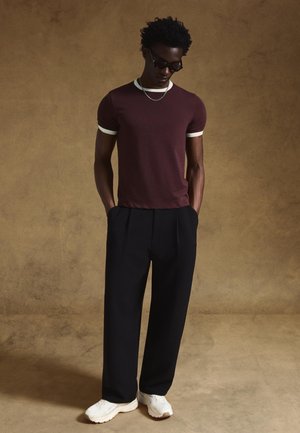 T-shirt color bordeaux in cotone con rifiniture bianche, abbinata a pantaloni neri plissettati e sneaker bianche, modella con occhiali da sole, in piedi con le mani nelle tasche.