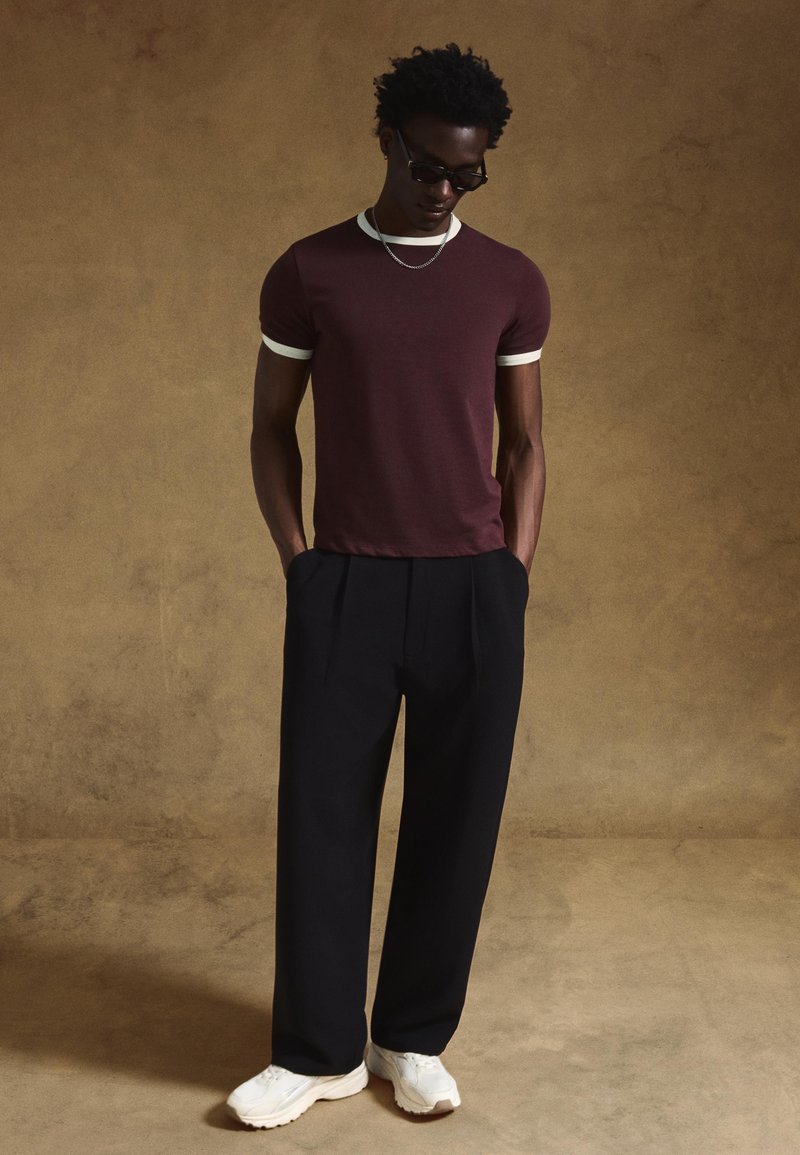 Bordo bombažna majica z belim robom, kombinirana s črnimi nagubani hlačami in belimi supergami, model nosi sončna očala, stoji z rokami v žepih.