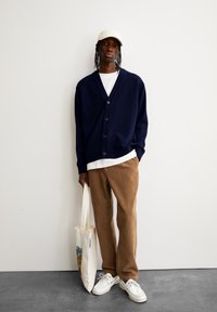 Cardigan bleu marine à cinq boutons, t-shirt blanc en dessous, pantalon en velours côtelé beige, chaussures blanches et casquette de baseball crème. Tiens un tote bag.