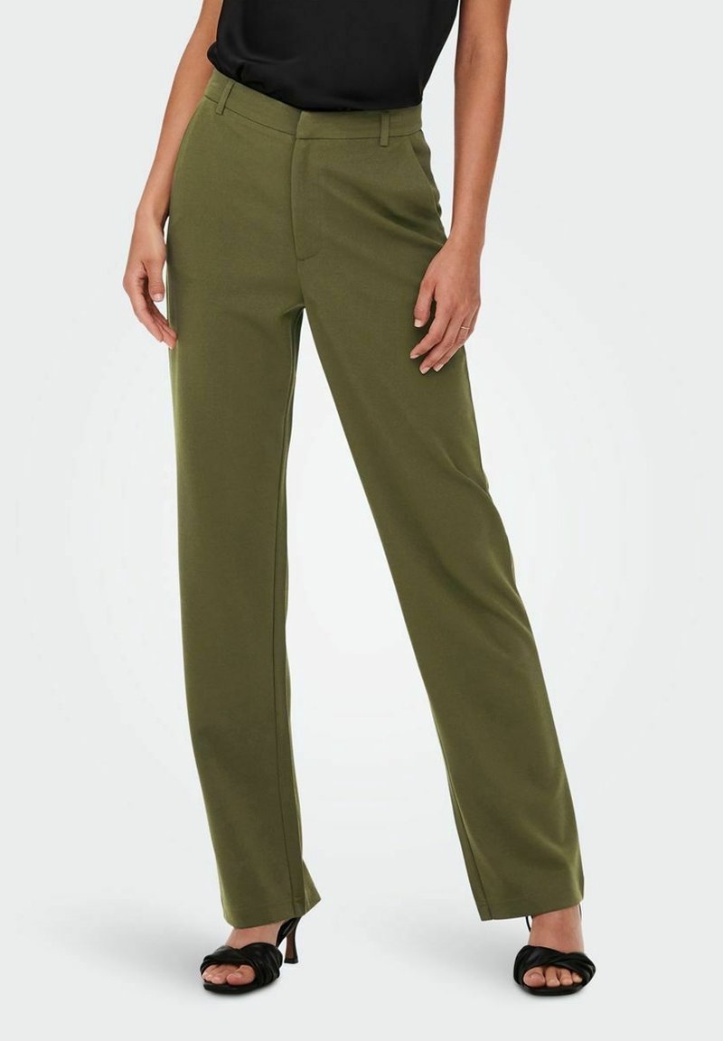 Žena v olivově zelených rovné kalhoty a černých sandálech na podpatku s otevřenou špičkou, stojící na jednobarevném světlém pozadí.