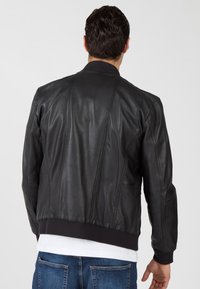 Zwarte leren bomberjack met een geribde kraag en manchetten, met verticale naden en zijzakken. Draagt over een wit shirt en jeans.