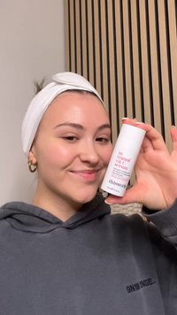 ThisWorks In Transit Vitamin C Serum in einem zylindrischen weißen Behälter mit pinkem Text, gehalten von einer Person in einem dunkelgrauen Hoodie.