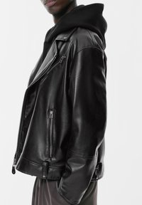 Crna kožna moto jakna s patentnim zatvaračem sprijeda, asimetričnim zakopčavanjem i patentnim džepovima s laganim sjajem. Nosena preko crne dukserice.