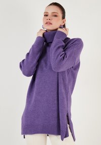Vijoličast oversize pulover s turtleneck ovratnikom in rebrastimi manšetami ter robom. Ima stranske razpoke in mehak teksturo za udobje.