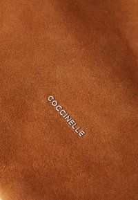 Une surface en daim marron au toucher lisse présente le logo argenté "Coccinelle" embossé dans une police élégante et moderne.