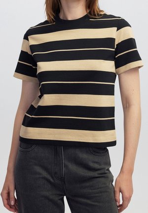 Kvinde iført en sort og beige kortærmet skjorte med horisontale striber og mørkegrå, højtaljede bukser med synlige lommer.