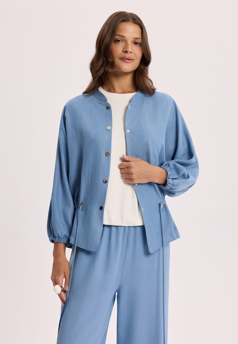 Kobieta ubrana w jasnoniebieską kurtkę zapinaną na guziki z elastycznymi mankietami, na białej bluzce i dopasowanych jasnoniebieskich spodniach na neutralnym tle.