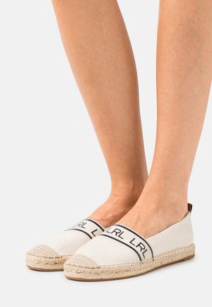 Espadrilles en toile blanche avec une marque noire sur le dessus, semelle en jute, et design plat. L'embout est légèrement pointu.