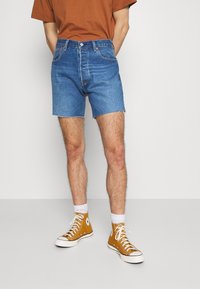 Jeansshorts i mellanblått, med rak passform och fransade kanter, kombinerat med gula högskurna sneakers och vita strumpor.