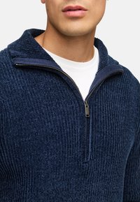 Mørkeblå fleece halv-zip sweater med ribbet tekstur og stående krave. Har en sølvfarvet lynlås og en hvid t-shirt under.