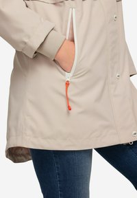 Ljusbeige vattentålig jacka med en framficka och orange snöre. Har en justerbar nederkant och dolda knäppningar.