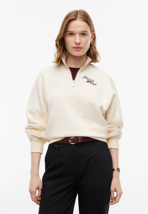 Ung kvinde iført en cremefarvet kvart-zip sweater med "Tommy Hilfiger" logo, sorte bukser, mørkerød bælte og guldkale, der står mod en hvid baggrund.